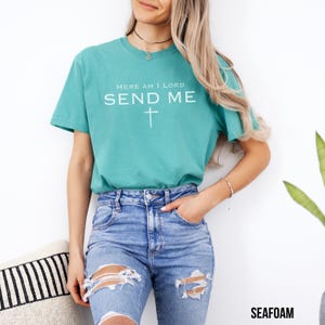 May include: A teal blue t-shirt with the text "Here am I Lord Send Me" and a white cross.