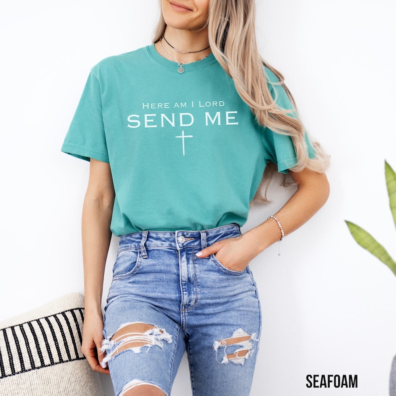 May include: A teal blue t-shirt with the text "Here am I Lord Send Me" and a white cross.