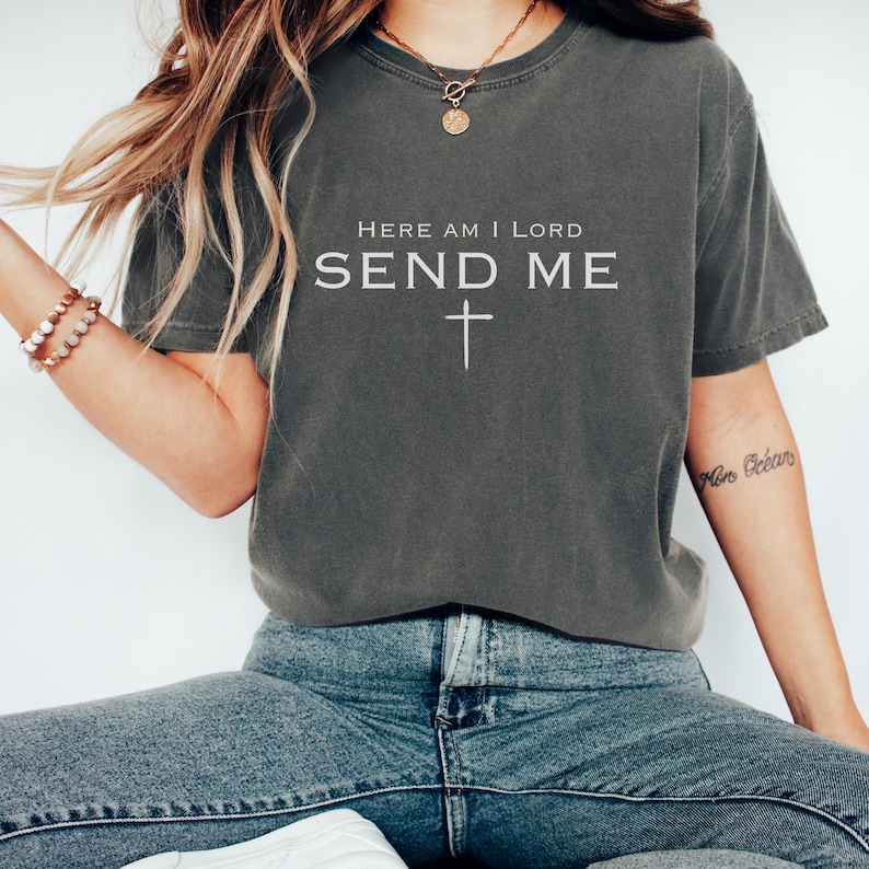 May include: A dark grey t-shirt with the text "Here am I Lord Send Me" and a cross underneath. The shirt is being worn by a person with long brown hair and a necklace.