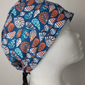 Puede incluir: Un gorro quirúrgico azul con un estampado repetido de conchas marinas en blanco, naranja y turquesa. El gorro tiene un cierre de lazo negro y se muestra en una cabeza de maniquí.