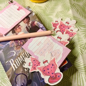 Könnte beinhalten: Nahaufnahme eines rosa und violetten Zaubertrankflaschen-Aufklebers mit Flügeln. Die Flasche ist mit rosa Flüssigkeit gefüllt und hat einen rosa Korken. Der Aufkleber befindet sich auf einem Buch mit dem Titel "The Romeo and Juliet  Dilemma".