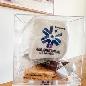 May include: Two clear plastic containers stacked on top of each other. The top container holds a white marshmallow with a blue and white logo that says "Europa Clipper" and "Avionics". The bottom container holds a similar marshmallow with the same logo. Both containers also contain graham crackers and a chocolate bar.