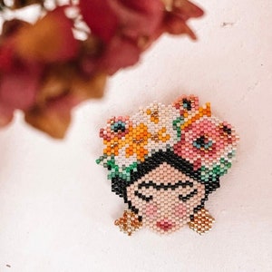 May include: A beaded portrait of Frida Kahlo with a floral crown. The portrait is made with colorful beads and features a detailed depiction of her face and hair. The crown is made of pink, yellow, and green beads.
