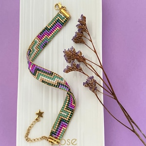 Peut inclure: Un bracelet de perles colorées avec un fermoir doré et un charm en forme d'étoile. Le bracelet présente un motif coloré de perles vertes, violettes et dorées. Il est présenté sur une assiette rectangulaire blanche avec un fond violet. Le texte "Rose Moustache" est visible.
