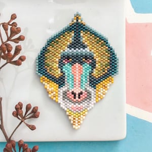 May include: A beaded mandrill monkey face with blue, green, yellow, and white beads. The monkey's face is beaded in a detailed and realistic style.