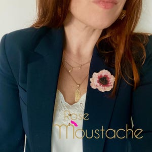 Pode incluir: Um blazer azul marinho com um broche de flor de contas em tons de rosa e preto. O broche é preso à lapela. Um colar de ouro com vários pingentes e o texto "Rose Moustache" é visível.