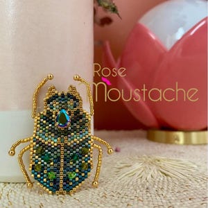 Puede incluir: Broche de escarabajo con cuentas, con patas y antenas de metal dorado. El cuerpo es un mosaico de cuentas azules, verdes y doradas, con una gema iridiscente. El texto "Rose Moustache" es visible.