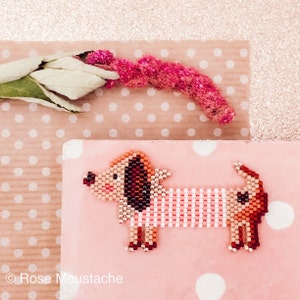 May include: A beaded dachshund wearing a pink and white striped shirt. The dachshund is brown, orange, and white. The dachshund is on a pink background with white polka dots.