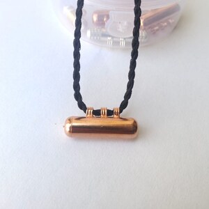 May include: A copper-colored cylindrical pendant hangs from a black braided cord. The pendant has three decorative rings around its center.