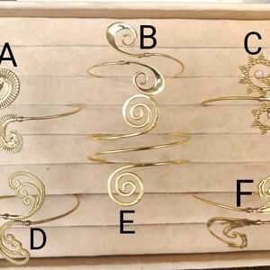 May include: Six gold-tone metal bracelets with different designs, including swirls, spirals, and a sunburst pattern. The bracelets are displayed in a white box with a brown interior.