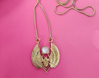 Könnte beinhalten: Eine goldene Halskette mit einem Skarabäus-Anhänger und einem weißen, schillernden Stein. Der Anhänger ist von Flügeln umgeben.