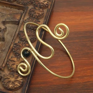 May include: A gold-colored arm cuff with a swirling design and a small, dark green stone. The cuff features two spiral accents and is displayed on a wooden surface with carved details.
