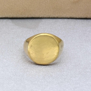 Puede incluir: Un anillo de sello de oro con una parte superior redonda y lisa. El anillo está hecho de un metal brillante y tiene un diseño simple.