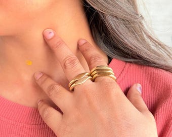 May include: Gold-colored rings with a leaf-like design. The rings are stacked on the fingers of a person wearing a pink shirt. The rings have a smooth, polished finish and are a warm, golden color.
