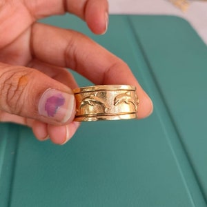 Puede incluir: Un anillo de color dorado con un diseño de delfines saltando fuera del agua. El anillo se lleva en un dedo.
