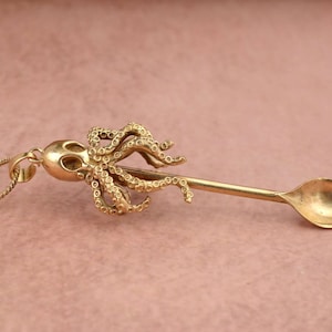 May include: A gold octopus spoon necklace. The octopus is detailed with textured tentacles and a skull-like head. The spoon is attached to the octopus's body and has a rounded bowl.