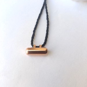 May include: A copper-colored pendant hangs from a black braided cord. The pendant is a small, cylindrical shape with rounded ends.
