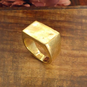 May include: A gold-toned signet ring featuring a smooth, rectangular flat top surface and a solid, thick band. The metal has a polished finish with subtle variations in texture across the surface, reflecting light across the top face.