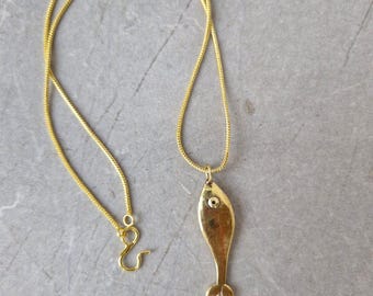 May include: A gold-colored necklace featuring a fish pendant. The pendant has a simple, stylized fish shape with a small circular detail representing an eye. The necklace chain is a fine, textured gold. The clasp is a hook and eye style.