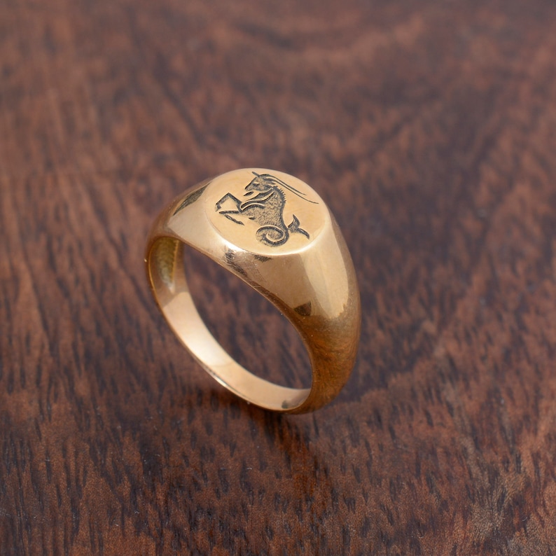 May include: A gold signet ring with a Capricorn zodiac symbol engraved on the top. The ring is oval shaped and has a smooth, polished finish.