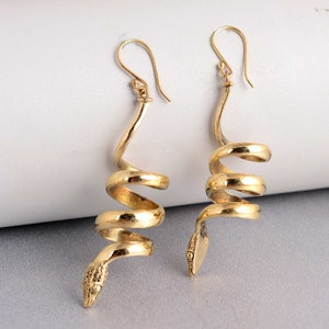 Puede incluir: Un par de pendientes colgantes de serpiente de color dorado. Los pendientes están hechos de un metal en espiral que se asemeja a una serpiente, con una cabeza de serpiente en la parte inferior.