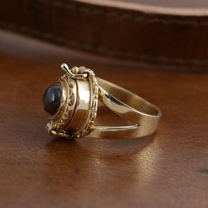 May include: A gold ring with a dark, round gemstone. The ring has an ornate, detailed setting with a hinged compartment. The band is split at the top, and the ring is displayed on a wooden surface.