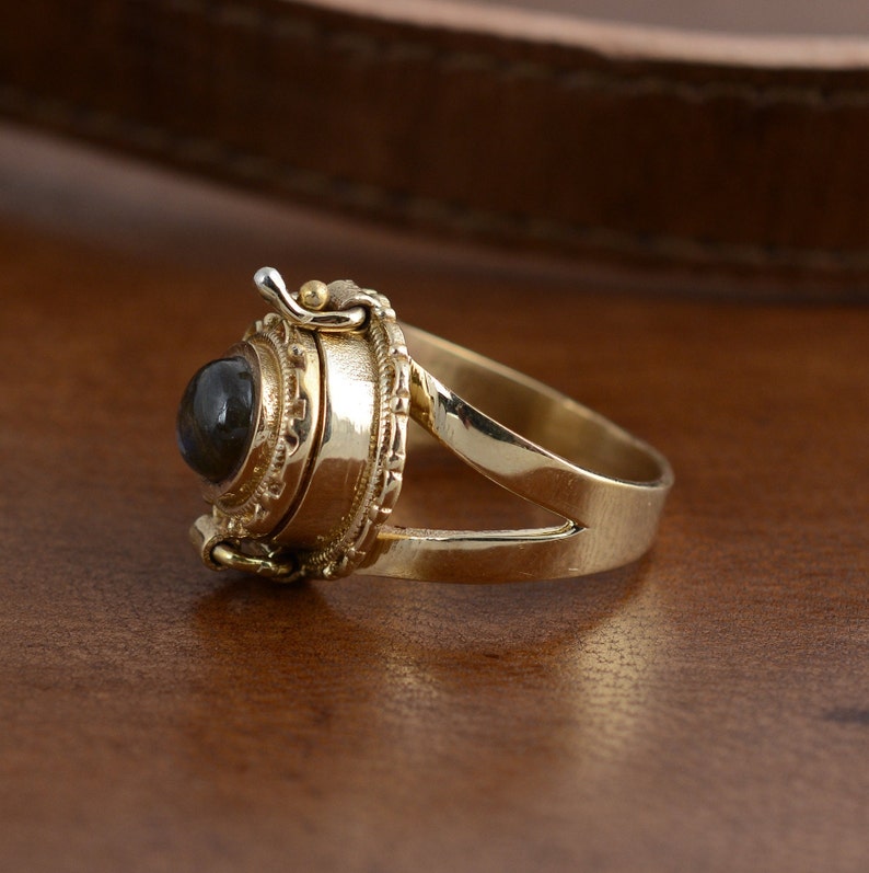 May include: A gold ring with a dark, round gemstone. The ring has an ornate, detailed setting with a hinged compartment. The band is split at the top, and the ring is displayed on a wooden surface.