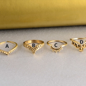 Peut inclure: Quatre bagues dorées de différents modèles sont présentées. La bague A a une forme en V avec de petites perles. La bague B a une forme en V similaire avec un motif de perles. La bague C est un demi-cercle avec de petites perles. La bague D a un design ajouré décoratif.
