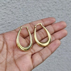 Peut inclure: Une paire de boucles d'oreilles créoles ovales dorées. Les boucles d'oreilles ont une surface lisse et polie et une forme de goutte d'eau classique. Les boucles d'oreilles sont présentées sur un fond gris clair.