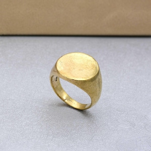 May include: A gold-toned signet ring with a smooth, circular flat top surface and a thick, solid band. The metal has a slightly weathered, matte finish with visible texture across the surface, showcasing a simple, unadorned design.