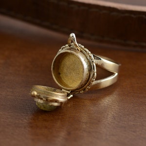 May include: A gold-colored locket ring with an open compartment. The ring has an ornate design around the compartment and a split band. The locket is open, revealing a small, light green stone inside.