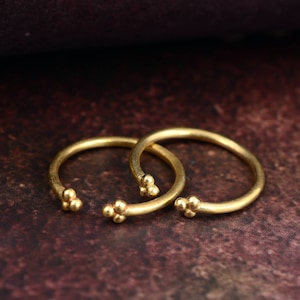 Puede incluir: Dos pendientes de aro abiertos de color dorado. Cada pendiente presenta un grupo de tres pequeñas cuentas redondas en el extremo abierto. Los pendientes se muestran sobre un fondo oscuro y texturizado, mostrando su diseño simple pero elegante.