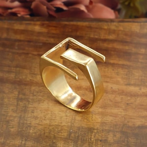 May include: A gold-toned ring featuring an open, geometric design with a wide, flat band that extends into two distinct, angular arms. The metal surface has a polished, reflective finish, showcasing a sharp, architectural silhouette with clean lines.