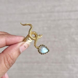May include: A gold-colored snake ring with a pear-shaped, light blue gemstone. The ring's design features a coiled snake wrapping around the finger, with the gemstone at the end of the snake's tail. The ring is held by a person's hand.
