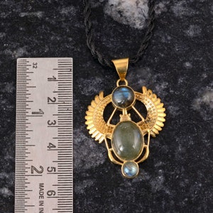 Op de afbeelding: Gouden hangerketting met een centrale ovale groene edelsteen en twee kleinere blauwe edelstenen. De hanger heeft een sierlijk ontwerp en een zwart koord. De hanger is ongeveer 5 cm lang, zoals te zien is op de liniaal.