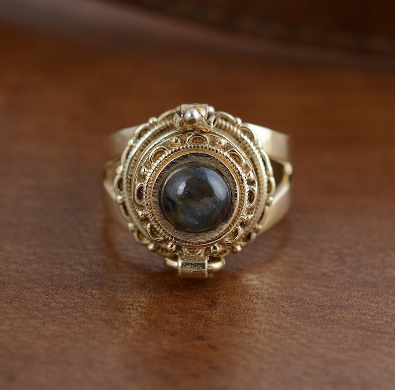 May include: A gold-colored ring with an ornate, detailed design. The ring features a central, dark-colored stone, surrounded by intricate filigree and decorative elements. The ring's design is reminiscent of vintage or antique jewelry.