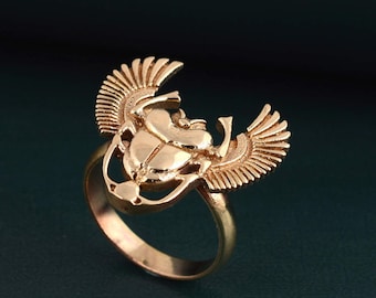 May include: A gold-toned ring featuring a detailed scarab beetle design with wings. The ring is made of metal and has a smooth, polished finish.