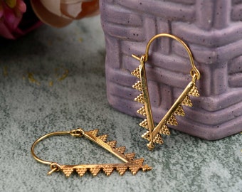 Peut inclure: Boucles d'oreilles créoles dorées avec un design en forme de V. Les boucles d'oreilles sont décorées de petits triangles.
