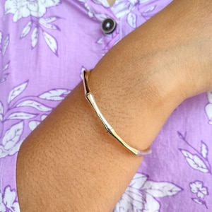 Op de afbeelding: Een gouden armband met een bamboe-ontwerp. De armband zit om een pols.