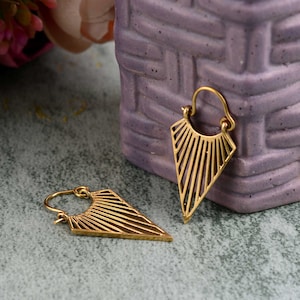 May include: A pair of gold-coloured earrings with a geometric design. Each earring has a triangular shape with parallel lines radiating from the top. The earrings are on a textured surface with a purple object in the background.