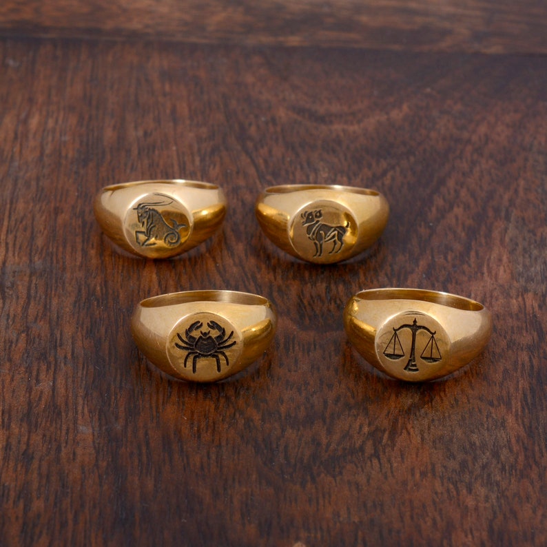 May include: Four gold signet rings with different zodiac symbols engraved on the top. The symbols are Capricorn, Aries, Cancer, and Libra.