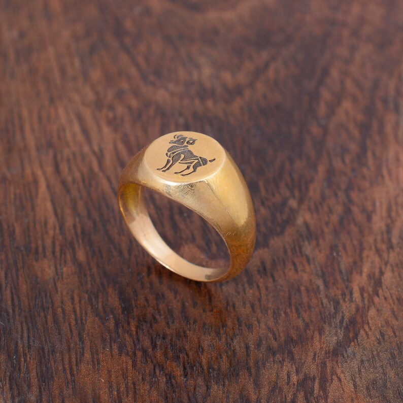 May include: A gold signet ring with a raised, round, engraved design of an Aries zodiac symbol.