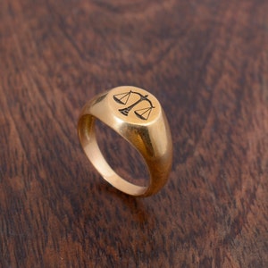 May include: A gold signet ring with a scale of justice design engraved on the top.