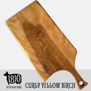 May include: A large, rectangular cutting board made of curly yellow birch wood with a handle. The board has a natural wood finish and a unique grain pattern. The board is engraved with the text "1810 BY DESIGN BARN".