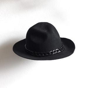 May include: A black fedora-style hat with a wide brim and a decorative chain band. The hat is made of a soft material and is suitable for various occasions. The hat is a classic accessory.
