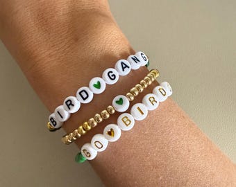 May include: Three beaded bracelets with the words "Bird Gang", "Go", and "Bird" spelled out in white beads with black and gold lettering. Each bracelet has a small green heart charm.