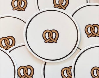 Op de afbeelding: Een set van zes witte onderzetters met een zwarte rand en een bruin pretzel-ontwerp in het midden.