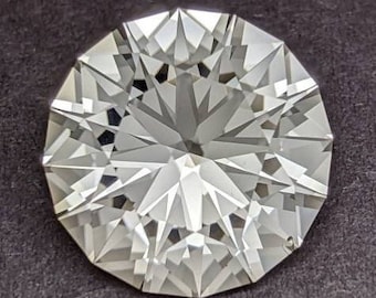 Puede incluir: Una gema redonda, transparente y facetada con un patrón de estrella. La gema está colocada sobre un fondo gris oscuro.