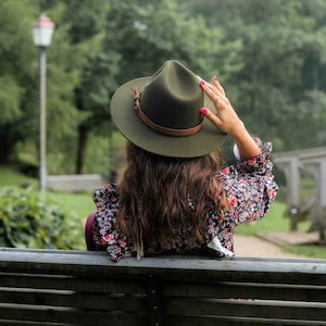 Può includere: Una donna con una blusa a stampa floreale e un cappello fedora verde scuro è seduta su una panchina di legno. La panchina si trova in un parco con alberi verdi sullo sfondo.