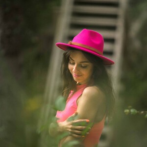 Puede incluir: Una mujer con un sombrero rosa y un top de tirantes coral. Est&aacute; de pie frente a una escalera de madera y vegetaci&oacute;n verde.